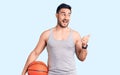 Young handsome man holding basketball ball pointing thumb up to the side smiling happy with open mouth Royalty Free Stock Photo