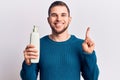 Young handsome man holding bottle of cream smiling with an idea or question pointing finger with happy face, number one Royalty Free Stock Photo