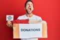 Young handsome man holding donations box and we need you paper angry and mad screaming frustrated and furious, shouting with anger Royalty Free Stock Photo