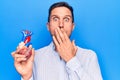 Young handsome man holding heart organ with veins and arteries over blule background covering mouth with hand, shocked and afraid