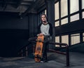 Young handsome man wearing shirt and hat holding his board while sitting on a grind rail in skatepark indoors