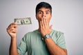 Young handsome man holding one dollar banknotes over isolated white background cover mouth with hand shocked with shame for Royalty Free Stock Photo