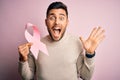 Young handsome man holding pink cancer ribbon symbol of support over isolated background very happy and excited, winner expression Royalty Free Stock Photo