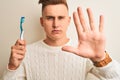 Young handsome man holding tooth brush standing over isolated white background with open hand doing stop sign with serious and Royalty Free Stock Photo