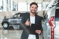 Young handsome man manager smiling holding brochure in his hand standing in dealership center near brand new SUV showing Royalty Free Stock Photo