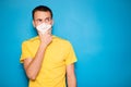 Young handsome man in medical mask and yellow t-shirt think how to dispose of problem isolated on blue background. Makes up an