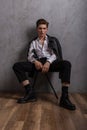Young handsome man model with trendy hairstyle in fashionable clothing in stylish boots sitting on a chair near a vintage wall in Royalty Free Stock Photo