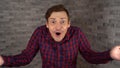 A young handsome man in plaid shirt opens his mouth and spreads his hands from surprise on a gray brick background.