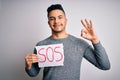 Young handsome man with problem holding banner with sos message over white background doing ok sign with fingers, excellent symbol