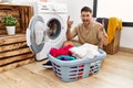 Young handsome man putting dirty laundry into washing machine shouting with crazy expression doing rock symbol with hands up Royalty Free Stock Photo