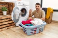 Young handsome man putting dirty laundry into washing machine showing and pointing up with fingers number four while smiling