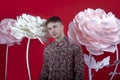 Young handsome man with rare little beard standing on bright red background between big white and pink artificial flowers. Royalty Free Stock Photo