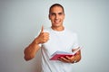 Young handsome man reading red book over white isolated background happy with big smile doing ok sign, thumb up with fingers, Royalty Free Stock Photo