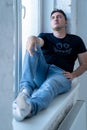 young handsome man with sad face, wearing jeans and t shirt is sitting on windowsill near the window, man looks through the window Royalty Free Stock Photo