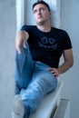 young handsome man with sad face, wearing jeans and t shirt is sitting on windowsill near the window, man looks through the window Royalty Free Stock Photo