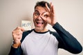 Young handsome man stressed holding reminder paper with help message with happy face smiling doing ok sign with hand on eye Royalty Free Stock Photo