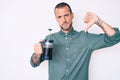 Young handsome man with tattoo holding french coffee maker with angry face, negative sign showing dislike with thumbs down, Royalty Free Stock Photo