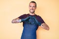 Young handsome man with tattoo wearing barber apron smiling in love showing heart symbol and shape with hands