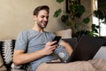 Young handsome man using his smartphone for online banking - sitting on sofa with laptop on couch Royalty Free Stock Photo