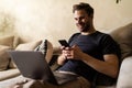 Young handsome man using his smartphone for online banking - sitting on sofa with laptop on couch Royalty Free Stock Photo