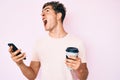 Young handsome man using smartphone holding take away coffee angry and mad screaming frustrated and furious, shouting with anger Royalty Free Stock Photo