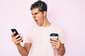 Young handsome man using smartphone holding take away coffee angry and mad screaming frustrated and furious, shouting with anger Royalty Free Stock Photo