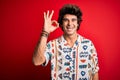 Young handsome man on vacation wearing summer shirt over isolated red background smiling positive doing ok sign with hand and Royalty Free Stock Photo