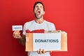 Young handsome man volunteer holding donations box with clothes angry and mad screaming frustrated and furious, shouting with Royalty Free Stock Photo