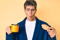 Young handsome man wearing bathrobe eating breakfast holding coffee and croissant making fish face with mouth and squinting eyes,