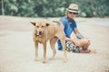 Young handsome man wearing blue t-shirt, hat and sunglasses, sitting on the beach with the dog in Thailand