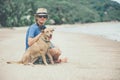 Young handsome man wearing blue t-shirt, hat and sunglasses, sitting on the beach with the dog in Thailand Royalty Free Stock Photo