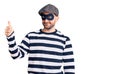Young handsome man wearing burglar mask looking proud, smiling doing thumbs up gesture to the side