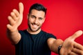 Young handsome man wearing casual black t-shirt standing over isolated red background looking at the camera smiling with open arms Royalty Free Stock Photo