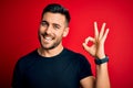 Young handsome man wearing casual black t-shirt standing over isolated red background smiling positive doing ok sign with hand and Royalty Free Stock Photo