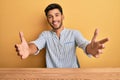 Young handsome man wearing casual clothes sitting on the table looking at the camera smiling with open arms for hug Royalty Free Stock Photo
