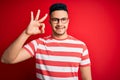 Young handsome man wearing casual striped t-shirt and glasses over isolated red background smiling positive doing ok sign with Royalty Free Stock Photo