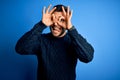 Young handsome man wearing casual sweater standing over isolated blue background doing ok gesture like binoculars sticking tongue Royalty Free Stock Photo