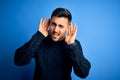 Young handsome man wearing casual sweater standing over isolated blue background Trying to hear both hands on ear gesture, curious Royalty Free Stock Photo