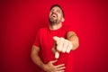 Young handsome man wearing casual t-shirt over red  background laughing at you, pointing finger to the camera with hand Royalty Free Stock Photo
