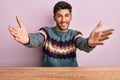 Young handsome man wearing casual winter sweater sitting on the table looking at the camera smiling with open arms for hug Royalty Free Stock Photo