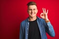 Young handsome man wearing denim shirt standing over isolated red background smiling positive doing ok sign with hand and fingers Royalty Free Stock Photo