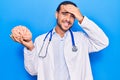 Young handsome man wearing doctor coat holding brain stressed and frustrated with hand on head, surprised and angry face Royalty Free Stock Photo