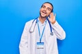 Young handsome man wearing doctor coat and stethoscope smiling with hand over ear listening and hearing to rumor or gossip Royalty Free Stock Photo