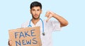 Young handsome man wearing doctor uniform holding fake news banner with angry face, negative sign showing dislike with thumbs