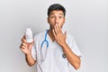 Young handsome man wearing doctor uniform holding presciption pills covering mouth with hand, shocked and afraid for mistake