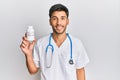 Young handsome man wearing doctor uniform holding presciption pills looking positive and happy standing and smiling with a