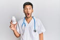 Young handsome man wearing doctor uniform holding presciption pills scared and amazed with open mouth for surprise, disbelief face