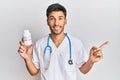 Young handsome man wearing doctor uniform holding presciption pills smiling happy pointing with hand and finger to the side