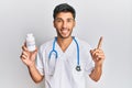Young handsome man wearing doctor uniform holding presciption pills smiling with an idea or question pointing finger with happy