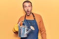 Young handsome man wearing gardener apron and gloves holding watering can scared and amazed with open mouth for surprise,